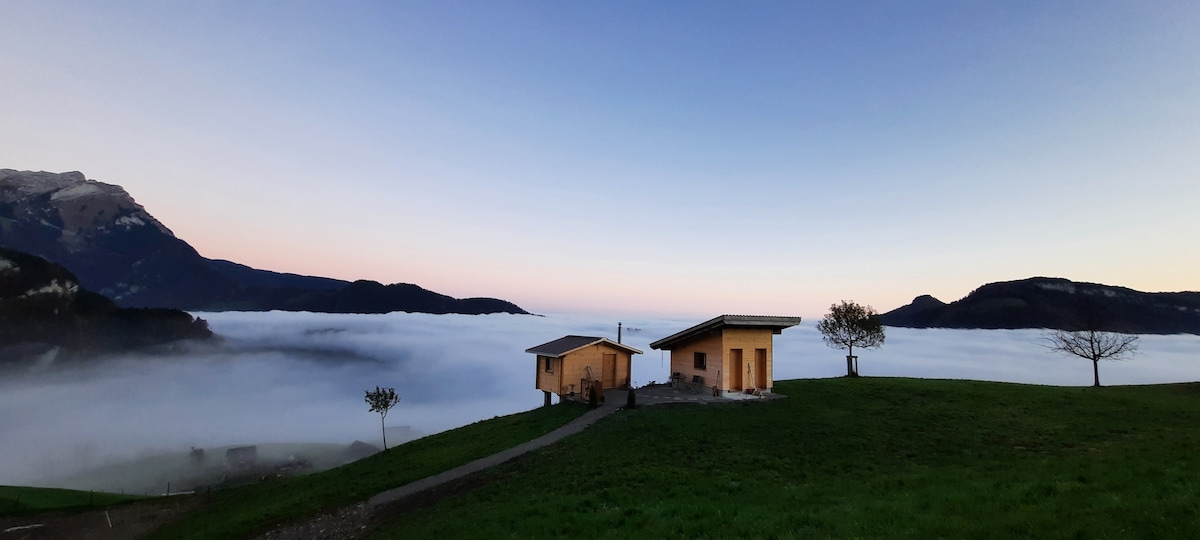 Two charming wooden houses are set against a backdrop of rolling fog and distant mountains. The scene captures the serene landscape at dawn, with soft pastel colors illuminating the sky. A path connects the houses, framed by lush green grass and scattered trees.