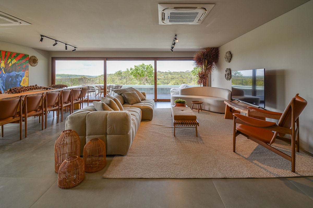 A spacious living area features large windows that frame views of the surrounding landscape. A comfortable sofa is positioned in the center, complemented by a sleek armchair. Woven baskets and a textured area rug enhance the warmth of the space, creating an inviting atmosphere.