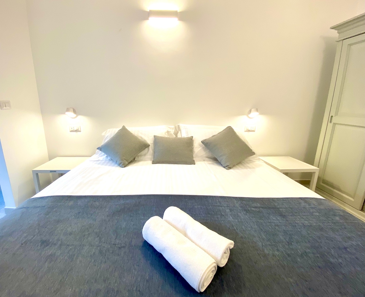 The bedroom features a spacious bed with white linens and decorative pillows against a light backdrop. Two bedside tables with wall-mounted lamps are positioned on either side. A neatly placed pair of rolled towels adds a touch of hospitality to the serene setting.
