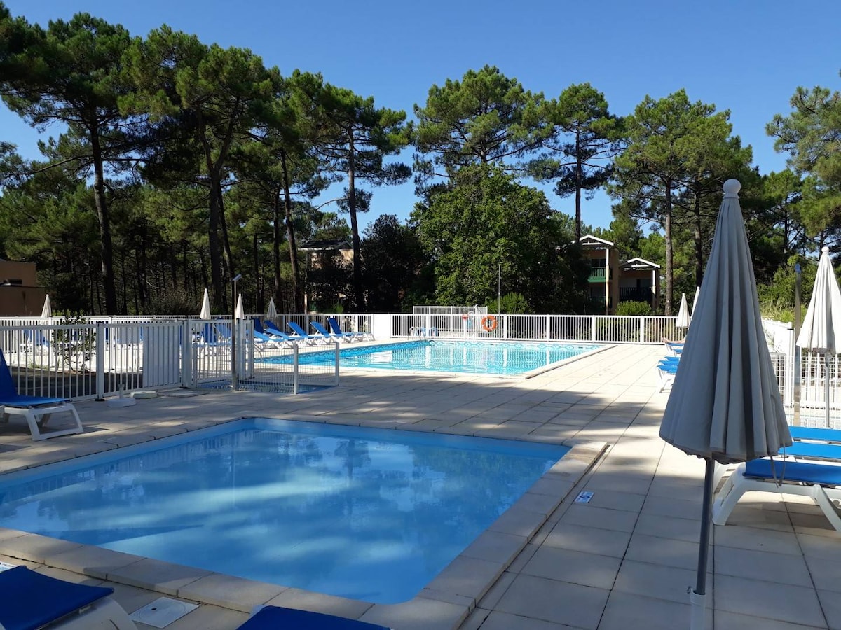 A communal swimming area is visible, featuring a large pool and a smaller, shallow pool. Sun loungers are arranged around the pool area, and distant pine trees provide shade. A white fence encloses the space, ensuring a serene atmosphere.