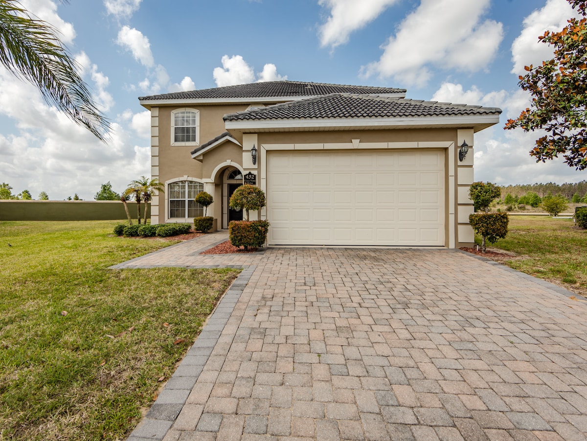 The exterior of the two-story home features a paved driveway leading to a two-car garage. Lush landscaping, including trimmed shrubs and small trees, surrounds the entrance, enhancing the welcoming appearance of the property. The sky is clear, with fluffy clouds adding depth to the scene.