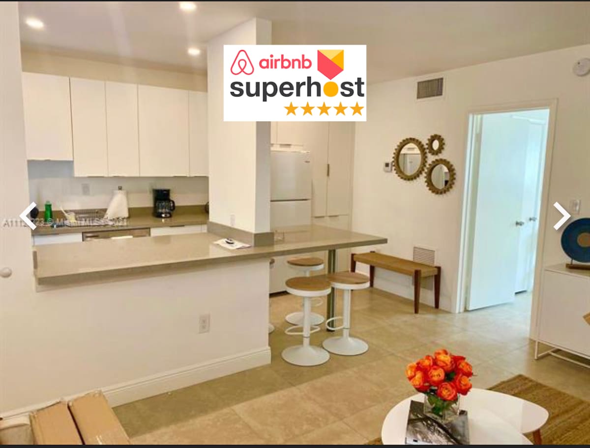 An open kitchen area is shown, featuring a light countertop and modern appliances. Two white stools are positioned at the breakfast bar. A vase with vibrant orange flowers sits on a neutral-colored rug in the living area, adding a touch of color and warmth.