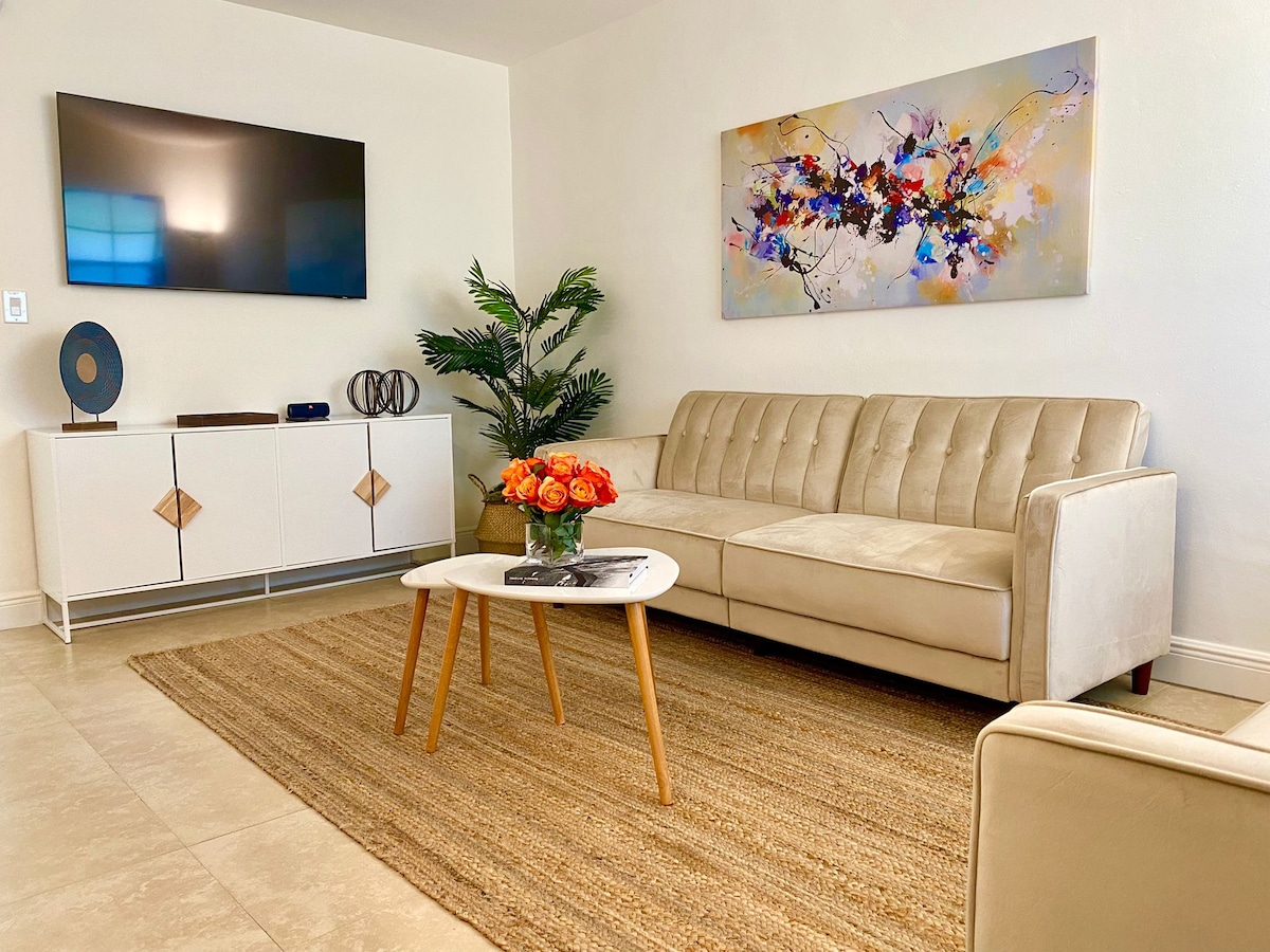 A bright and modern living area is presented, featuring a light-colored sofa paired with a round coffee table. A vibrant piece of wall art adds color to the space, while a tall plant brings a touch of greenery. A television is mounted on the wall, enhancing the room's functionality.