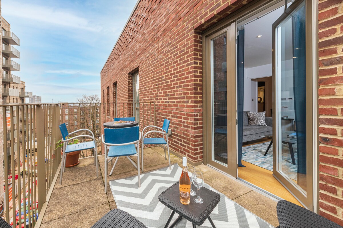 A private balcony is presented with outdoor seating, featuring two blue chairs and a small black table. The balcony offers a view of the surrounding area, with an open door leading to the interior space. A light-patterned rug adds texture to the outdoor setting.