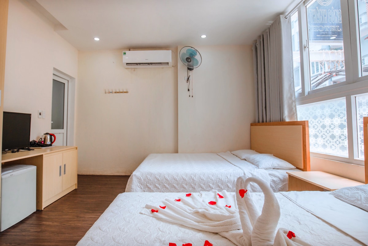 A bright room features two spacious beds with white linens adorned with red petals. Natural light fills the space through large windows, while a small television and a refrigerator are positioned along one wall. An air conditioning unit is installed for comfort.