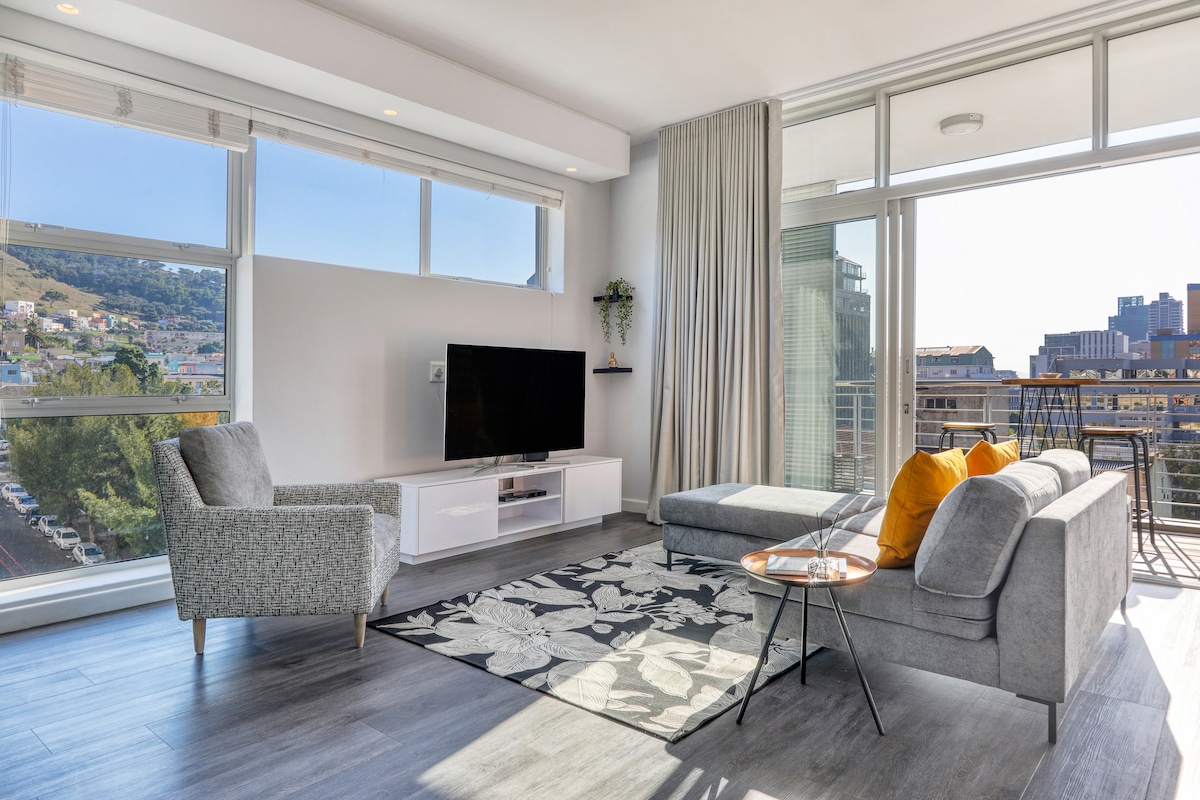 A modern living area features large windows providing ample natural light and views of the surrounding cityscape. A comfortable, light gray sectional sofa is complemented by a patterned area rug. A flat-screen television is mounted on a white media console, and a small table is positioned beside the sofa.