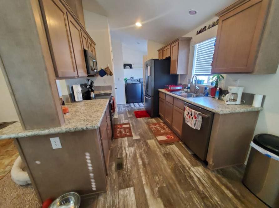 The kitchen features light wood cabinetry and granite countertops, providing ample workspace. A black refrigerator and microwave are visible, alongside a double sink. Decorative mats and a few kitchen essentials add warmth to the space. Natural light enters through a window, illuminating the tiled floor.