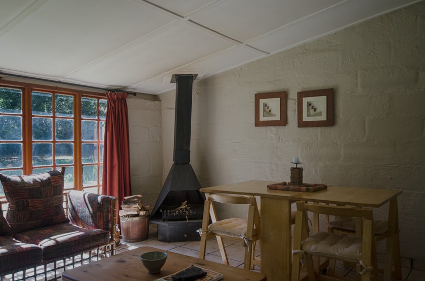 A comfortable interior space features a cozy seating area with a patterned armchair next to a fireplace. A simple wooden table with two chairs is positioned nearby, while natural light filters through large windows adorned with red curtains.
