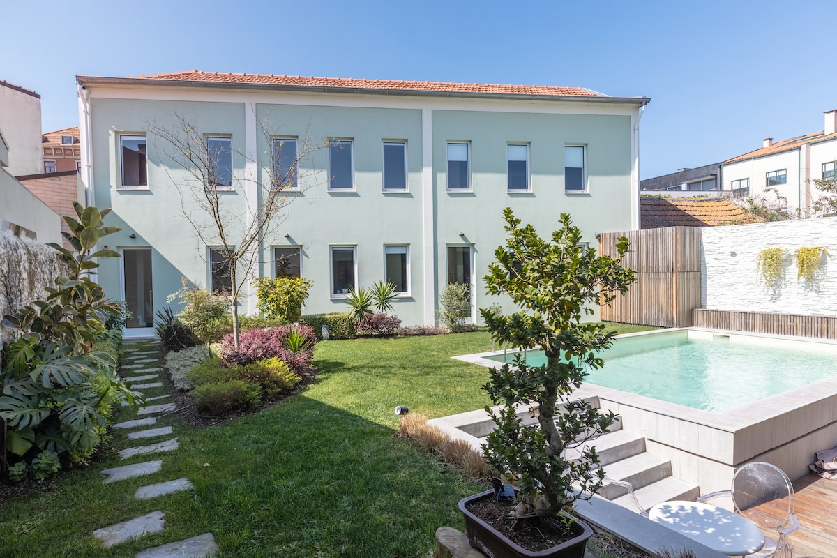 The exterior features a modern building with light green walls, showcasing multiple windows. A serene garden surrounds the space, accentuated by various plants and a neatly trimmed lawn. A pool is visible in the background, inviting relaxation in the sunshine.