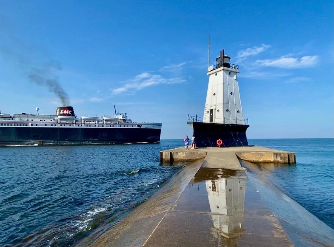 A view of a lighthouse situated on a pier, with calm waters extending toward the horizon. In the background, a large ship is seen navigating through the lake, accompanied by a clear blue sky. The pier's surface reflects the lighthouse and the ship.