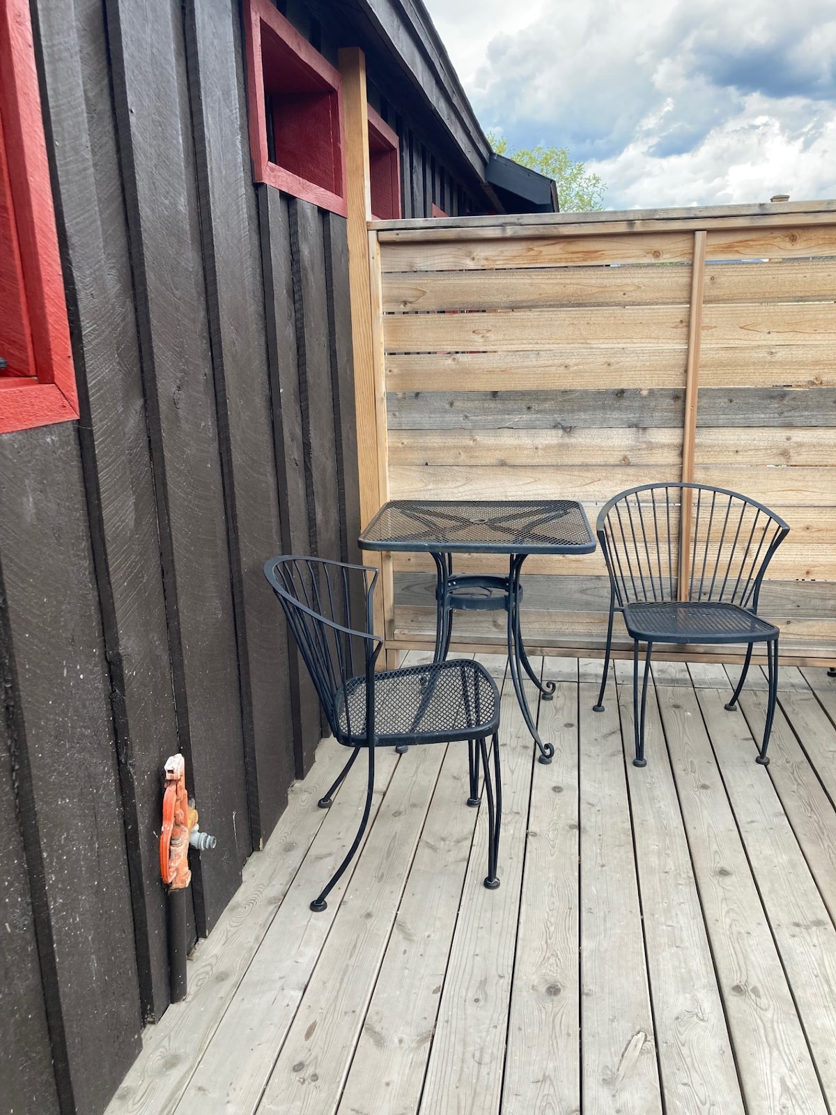 An outdoor porch area features a small black metal table accompanied by two matching chairs. The space is bordered by wooden planks and has red accent windows, creating a cozy and inviting spot for relaxation.
