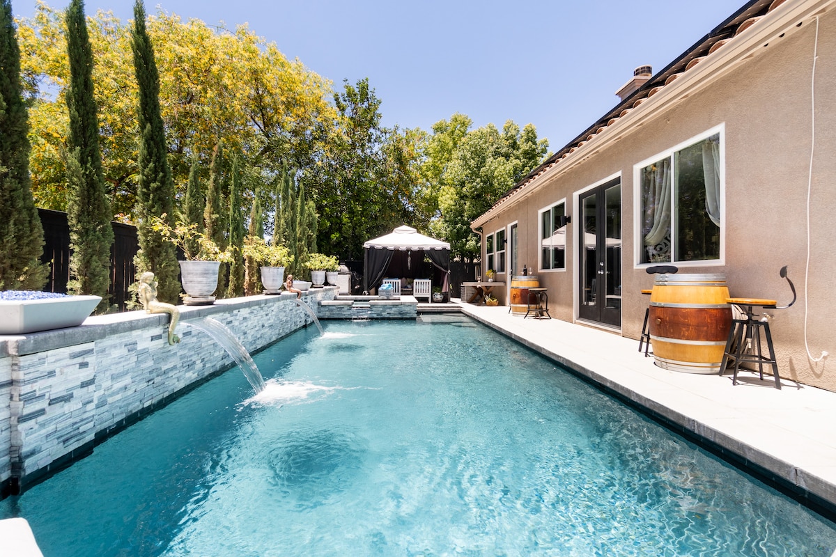 A serene outdoor space is highlighted by a sparkling pool with a waterfall feature. Sun loungers are positioned nearby, and a shaded cabana provides a relaxing retreat. Lush greenery surrounds the area, creating a tranquil atmosphere for guests to enjoy.