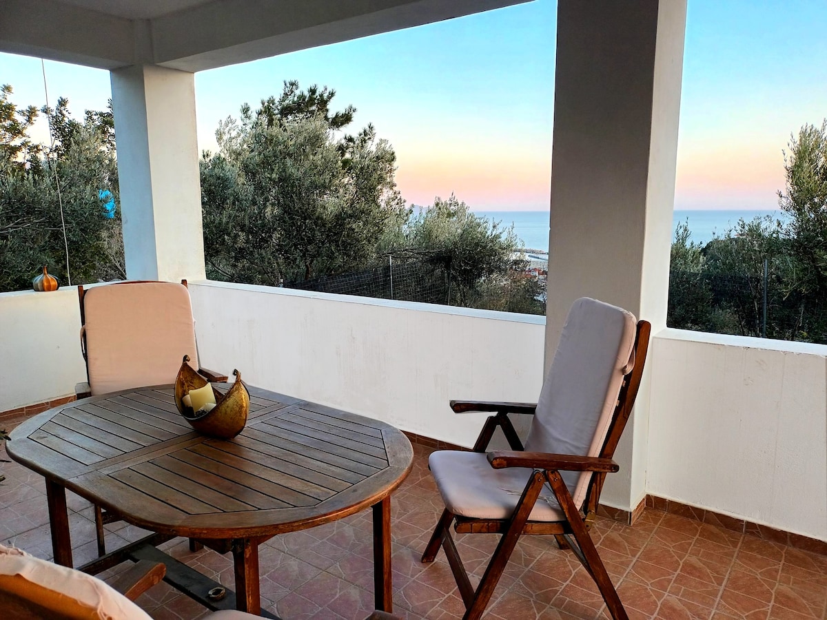 A spacious balcony features a wooden table surrounded by two comfortable chairs. A decorative bowl sits at the center of the table. The scenic view includes olive trees and the tranquil sea, illuminated by gentle hues from the evening sky.