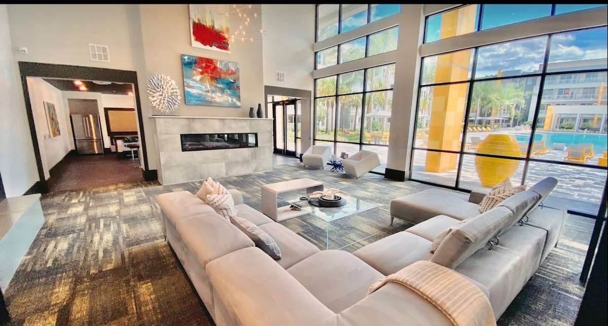 An open living space features a large sectional sofa facing a modern fireplace. Large windows allow natural light to flood the area, showcasing outdoor views. Artistic wall pieces and a stylish coffee table enhance the contemporary design of the space, creating an inviting atmosphere.
