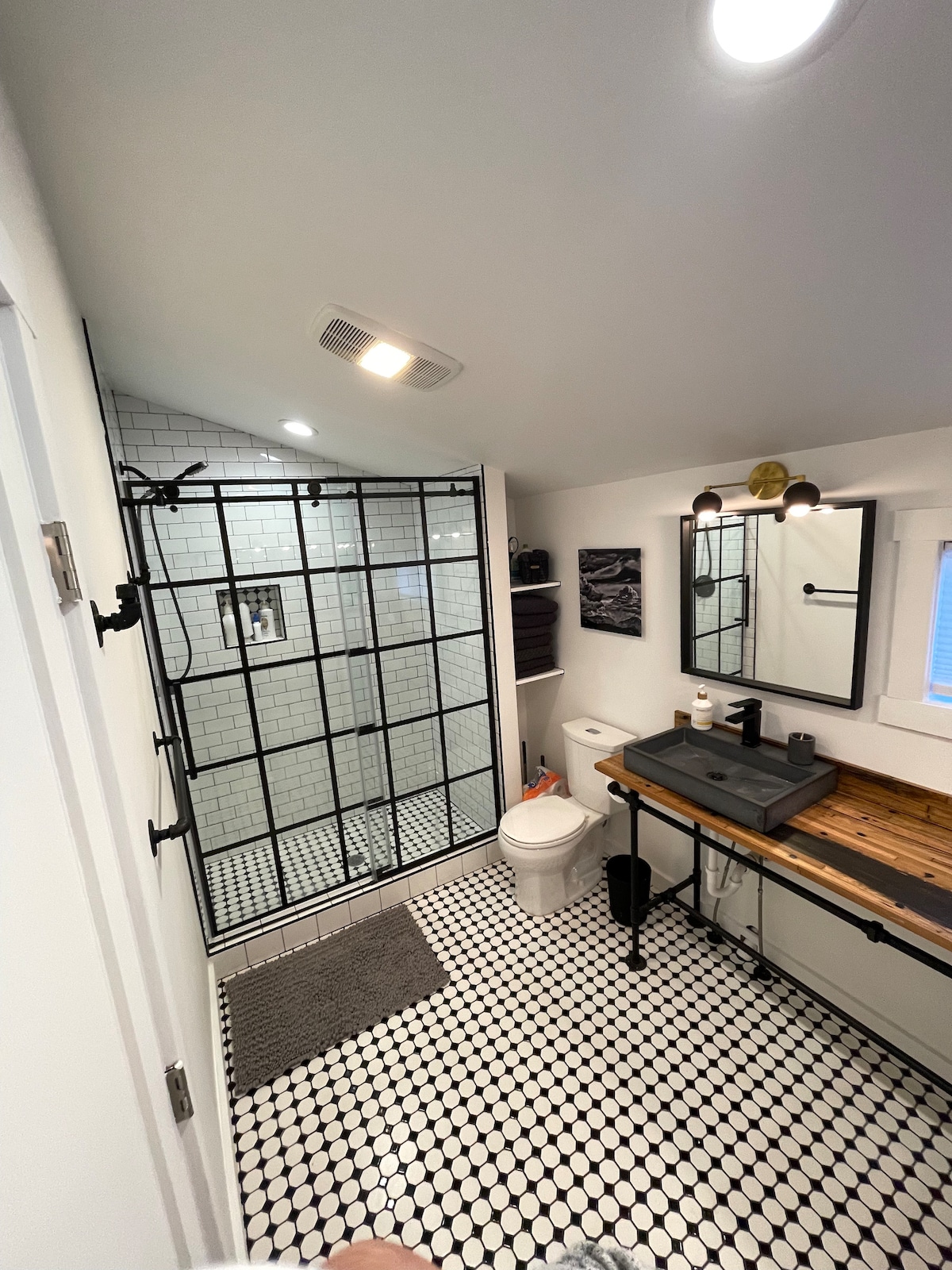A modern bathroom is presented, featuring a spacious walk-in shower with glass paneling. A sleek black vanity with a wooden countertop is positioned opposite a white toilet. The floor is covered with black and white patterned tiles, and framed artwork adds a touch of decor.