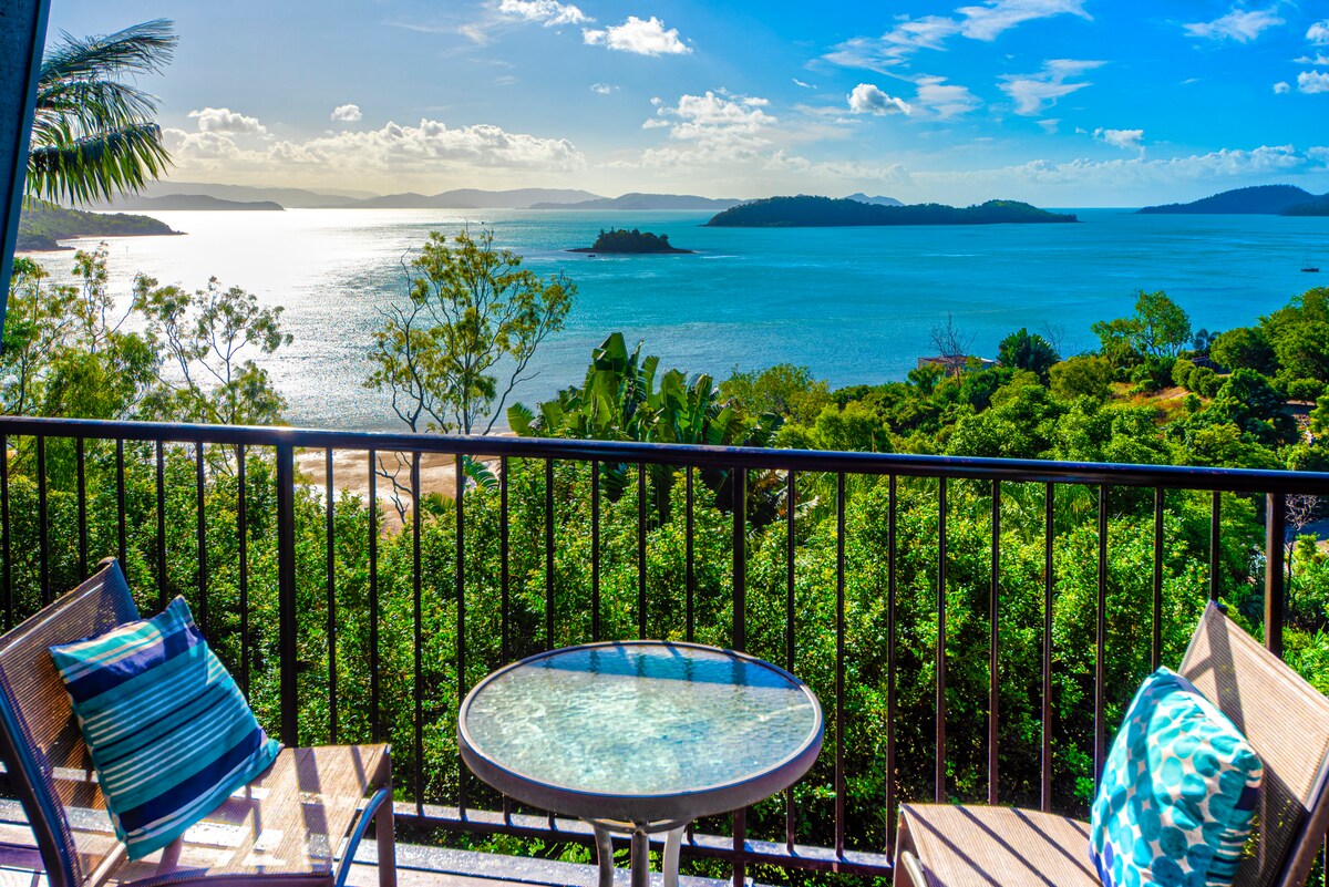 A scenic view of the Whitsunday Islands is captured from a balcony. Two outdoor chairs and a round table are visible, placed on a surface that reflects light. Lush greenery frames the landscape, and the azure waters stretch toward the horizon under a clear sky.