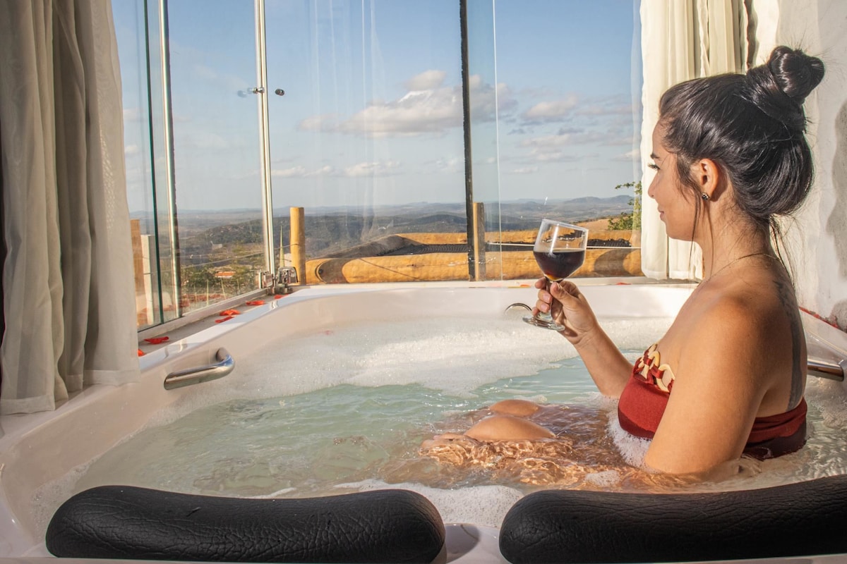 A spacious hot tub is situated by large glass windows that offer a scenic view of the surrounding mountains. Bubbles create a relaxing atmosphere, while a drink is held by a guest enjoying the serene setting.