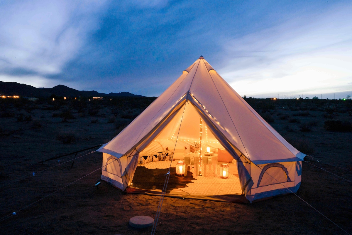 A spacious bell tent is set on the desert ground, softly illuminated by warm lanterns. Inside, comfortable seating and decorative elements create a cozy ambiance. The backdrop features a vast desert landscape under a twilight sky, enhancing the serene atmosphere.