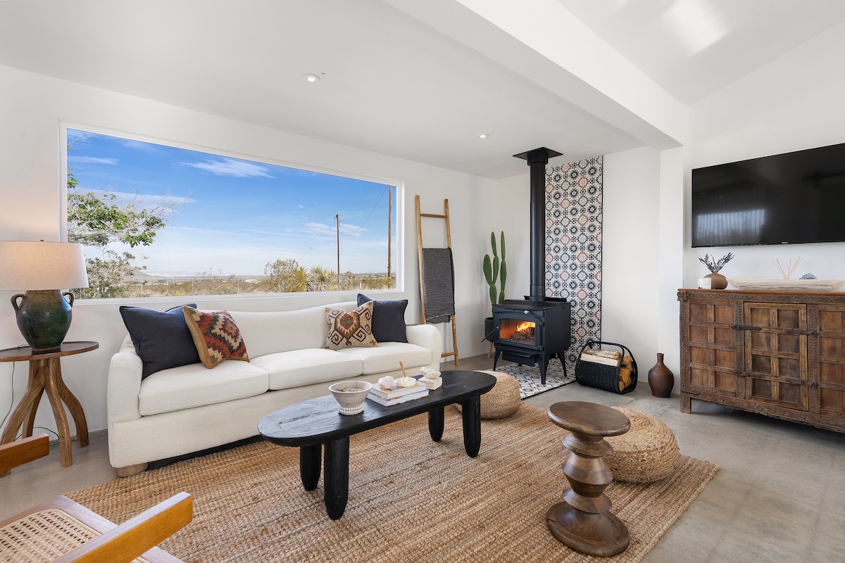 A spacious living area features a cozy white sofa accented with colorful cushions and an area rug. A wood-burning stove stands in the corner, while large windows provide ample natural light and views of the outdoors.