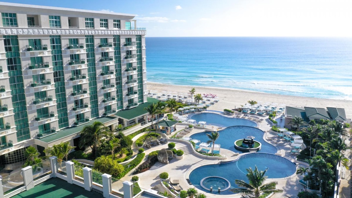 The image features a large resort building beside multiple elegantly designed infinity pools. Lush palm trees and lounge chairs surround the pools, while the sandy beach and the shimmering Caribbean Sea are visible in the background under a clear blue sky.