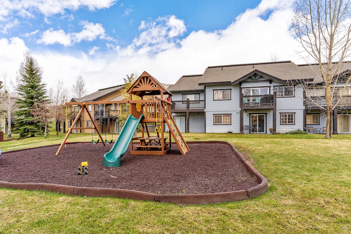 A shared playground is visible, featuring a wooden structure with a slide and swings, surrounded by a soft mulch surface. The townhome's spacious exterior includes multiple balconies, with grassy areas and trees in the background. The sky is partly cloudy, providing natural light to the scene.