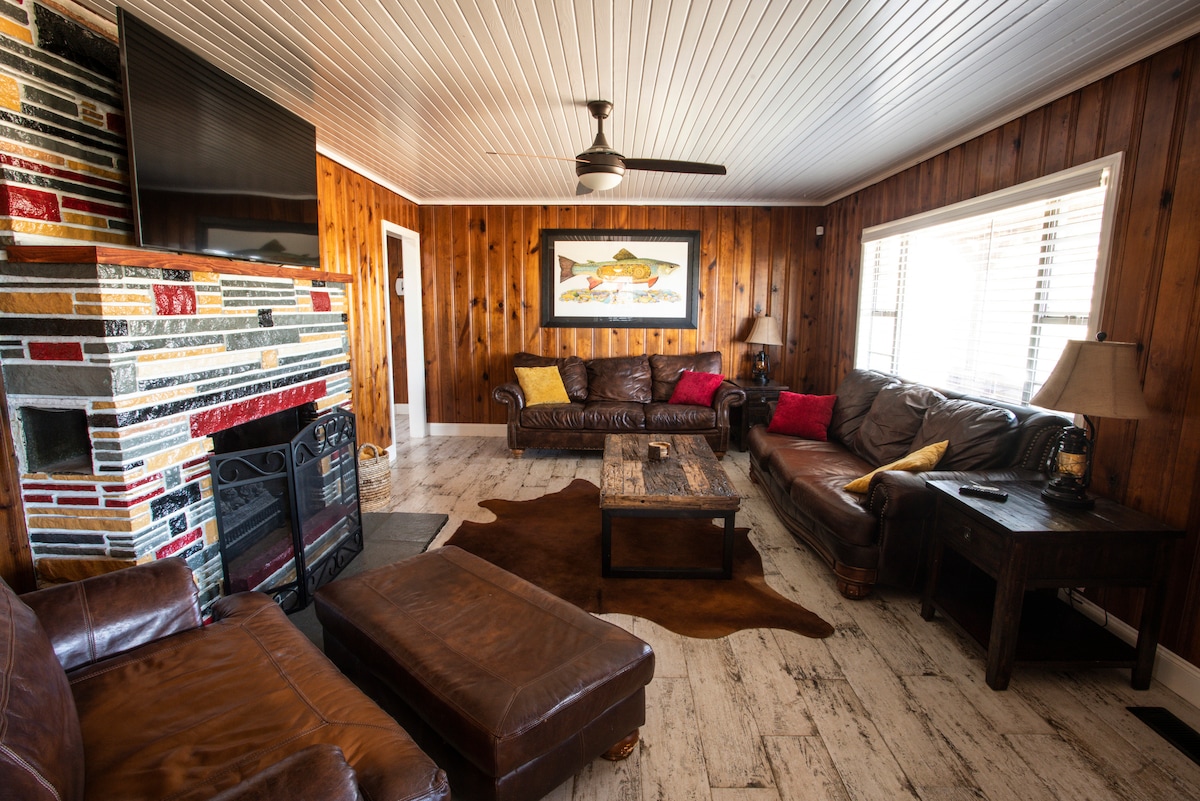 The living room features wood-panelled walls and a stone fireplace, complemented by a flat-screen TV. Leather seating arrangements, including a spacious couch and lounge chair, are arranged around a rustic coffee table. Soft lighting from table lamps enhances the cozy feel of the space.