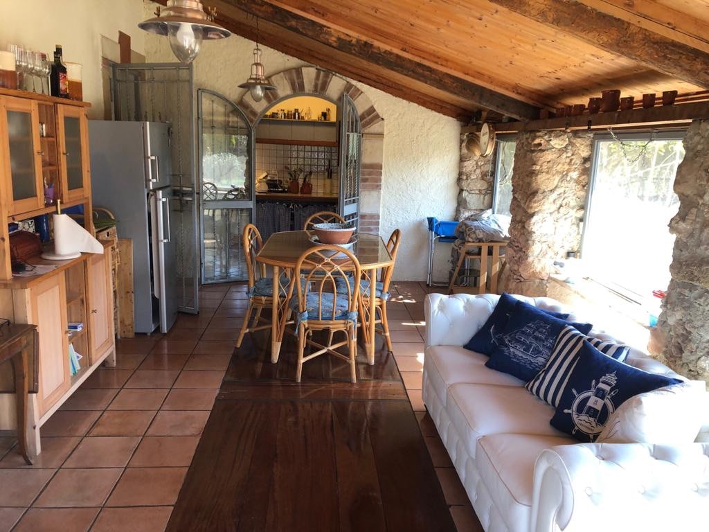 An open-plan living and dining area features a light-colored sofa and a wooden dining table surrounded by cane chairs. Exposed wooden beams add character to the high ceiling, while a stone wall provides rustic charm. Natural light fills the space through large windows.