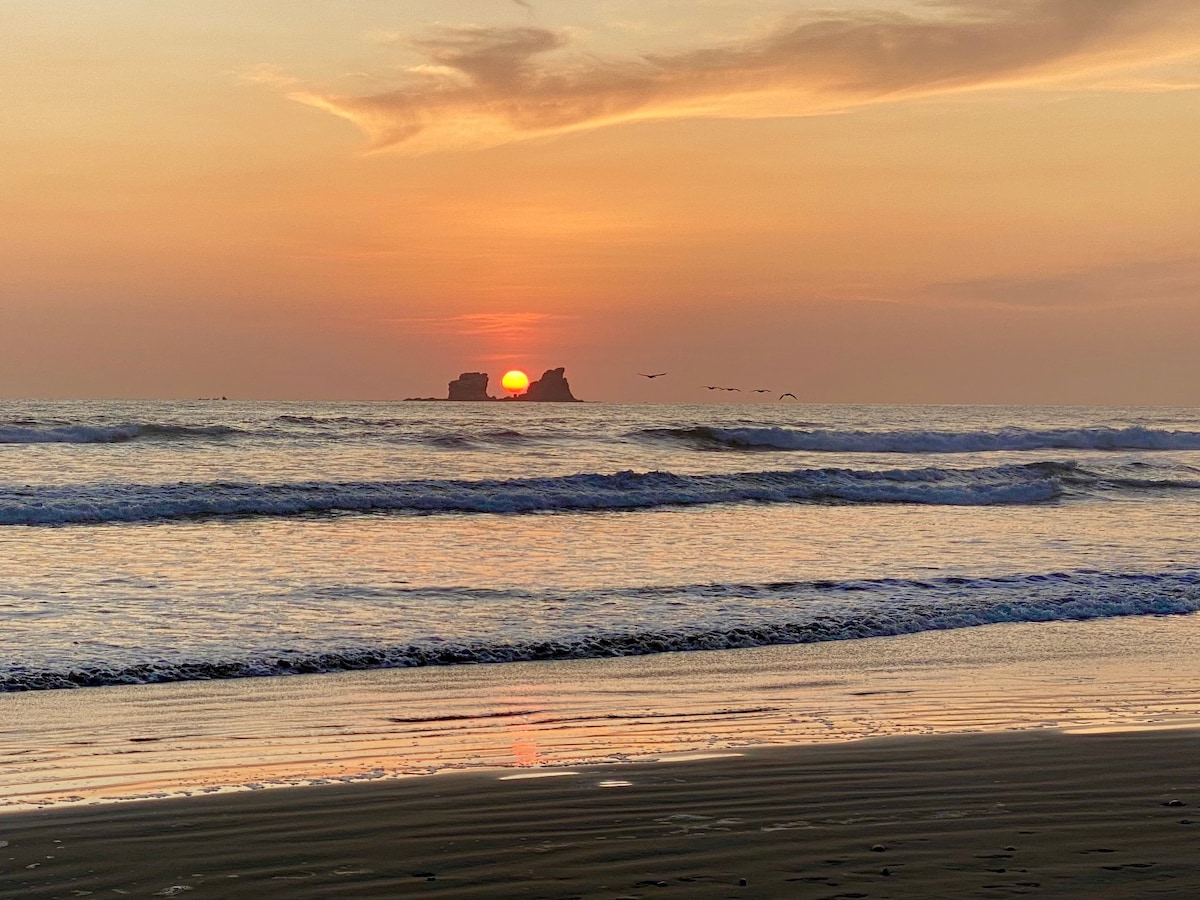 The sun sets over the ocean, casting warm hues of orange and pink across the sky. Waves gently lap at the shore, reflecting the colorful light. The silhouette of an islet is visible in the distance, while a few birds fly near the horizon.
