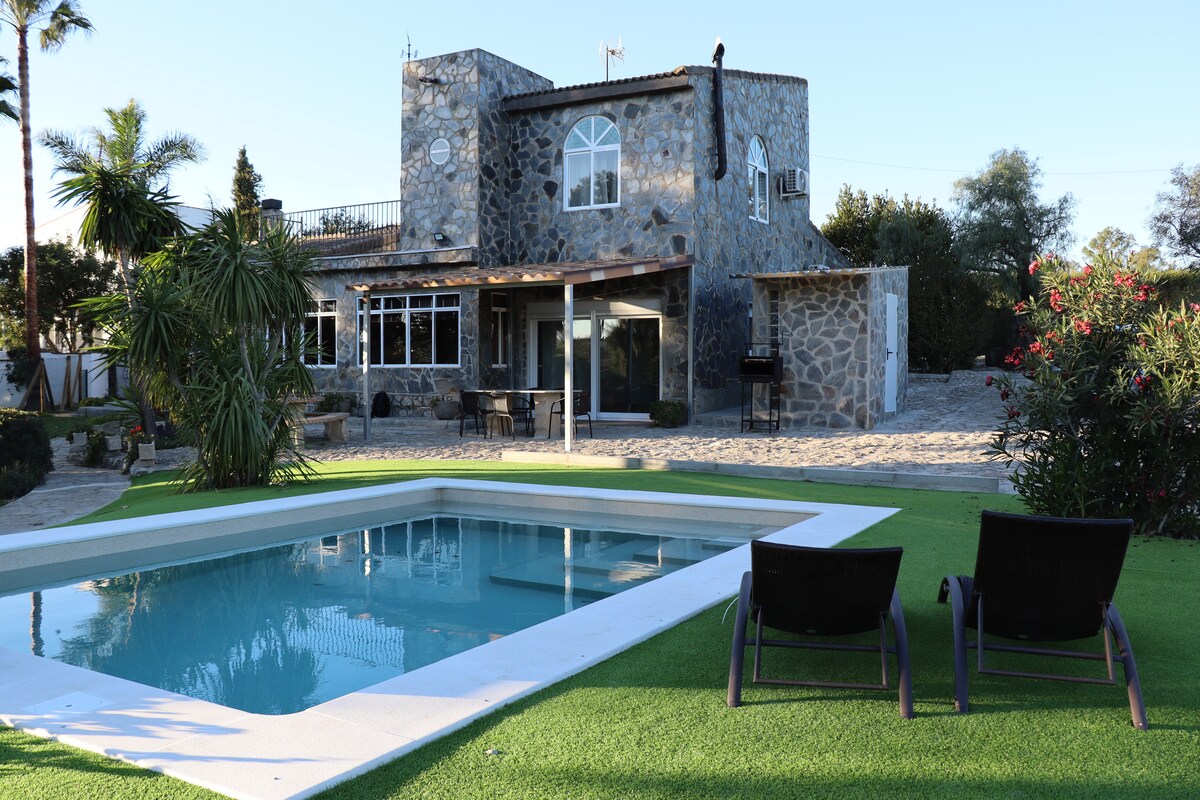 A stone-constructed house with large windows is set against a clear sky. In the foreground, a rectangular saltwater pool reflects the surroundings, with two lounge chairs positioned nearby on a grassy area. Lush greenery and flowering plants enhance the outdoor space.