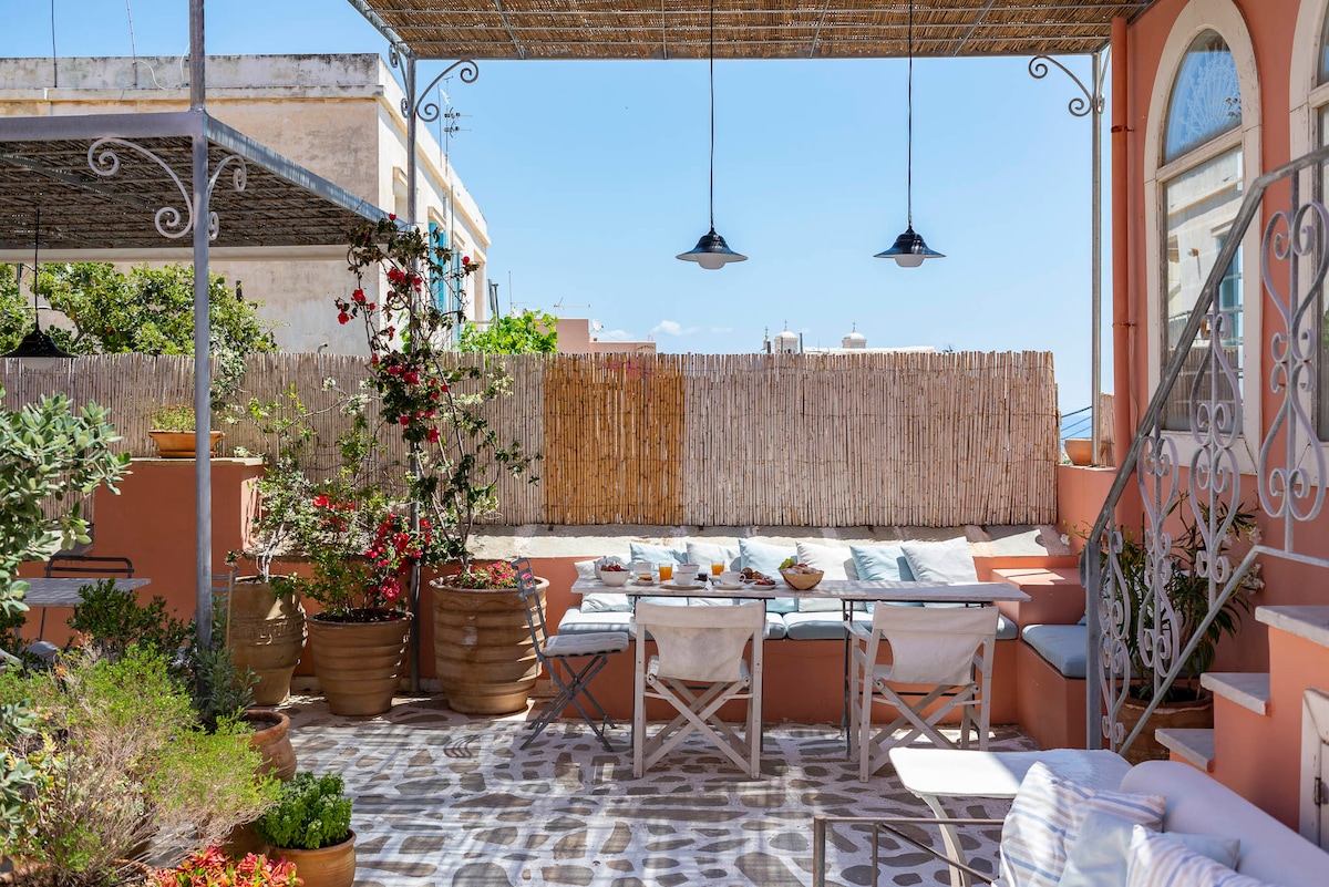 A charming outdoor terrace is showcased, featuring a shaded seating area with light-colored cushions and a low table set for breakfast. Potted plants and colorful flowers add vibrancy, while a bamboo screen provides privacy. The patterned flooring enhances the inviting atmosphere.