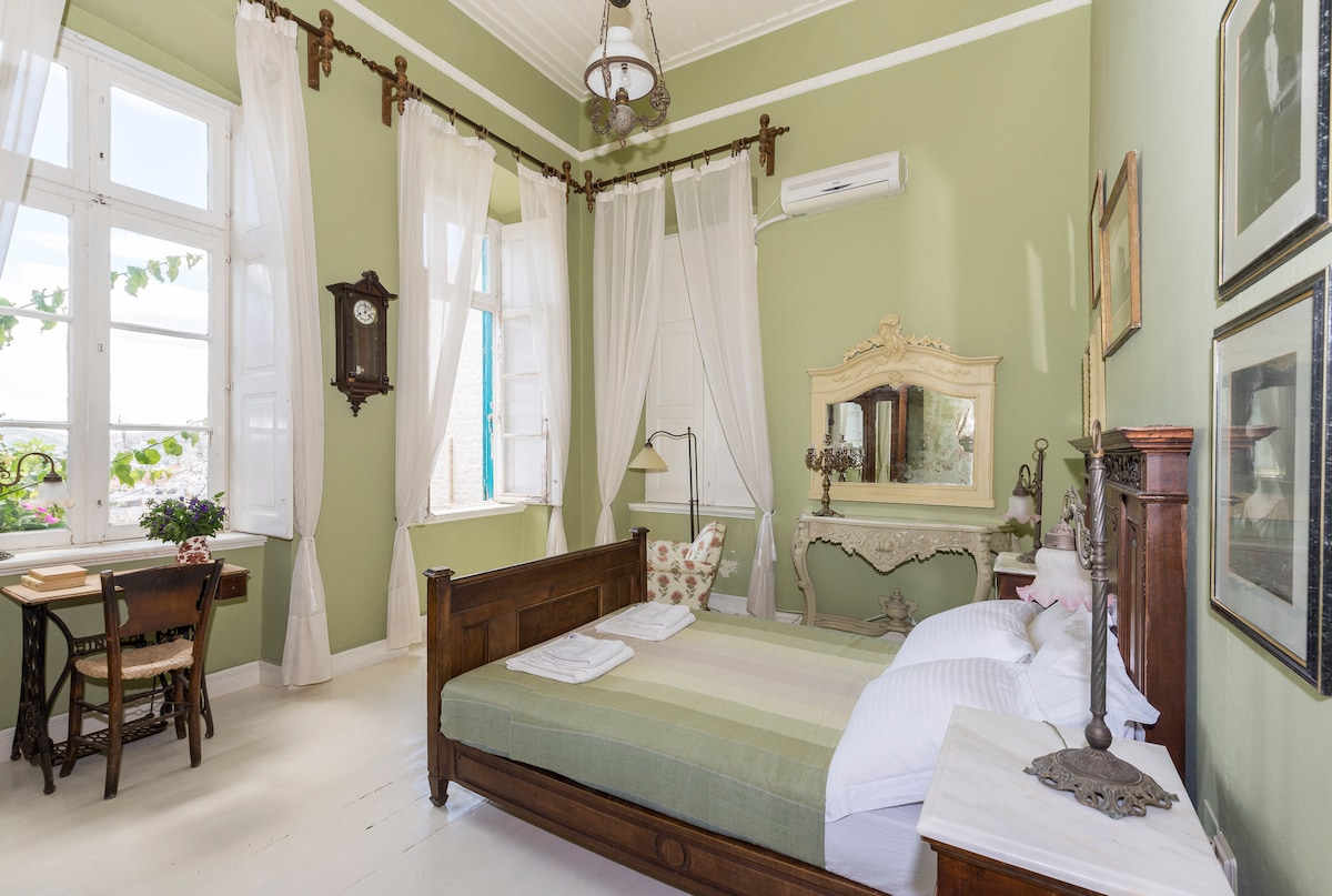 A serene bedroom is highlighted by soft green walls and large windows that allow natural light to fill the space. A spacious bed is adorned with neutral linens, while a vintage table and chairs are positioned by the window. Elegant decor adds to the room's charm.