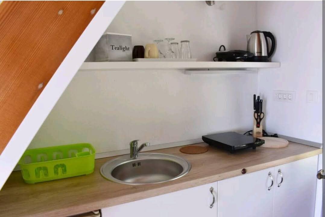 A compact kitchen area is featured, showcasing a stainless steel sink and a wooden countertop. Above, a shelf holds glassware and kitchen appliances, including a kettle and a griddle. An organized drawer with cooking utensils is visible, enhancing practicality.
