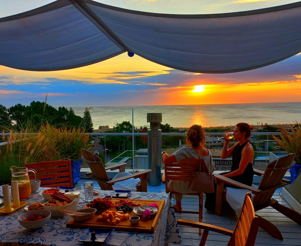 A terrace setting is captured at sunset, with a backdrop of a vibrant sky and ocean view. Two individuals are seated, enjoying a meal spread across a table adorned with various dishes. Comfortable wooden chairs provide a cozy atmosphere, while the awning offers shade.