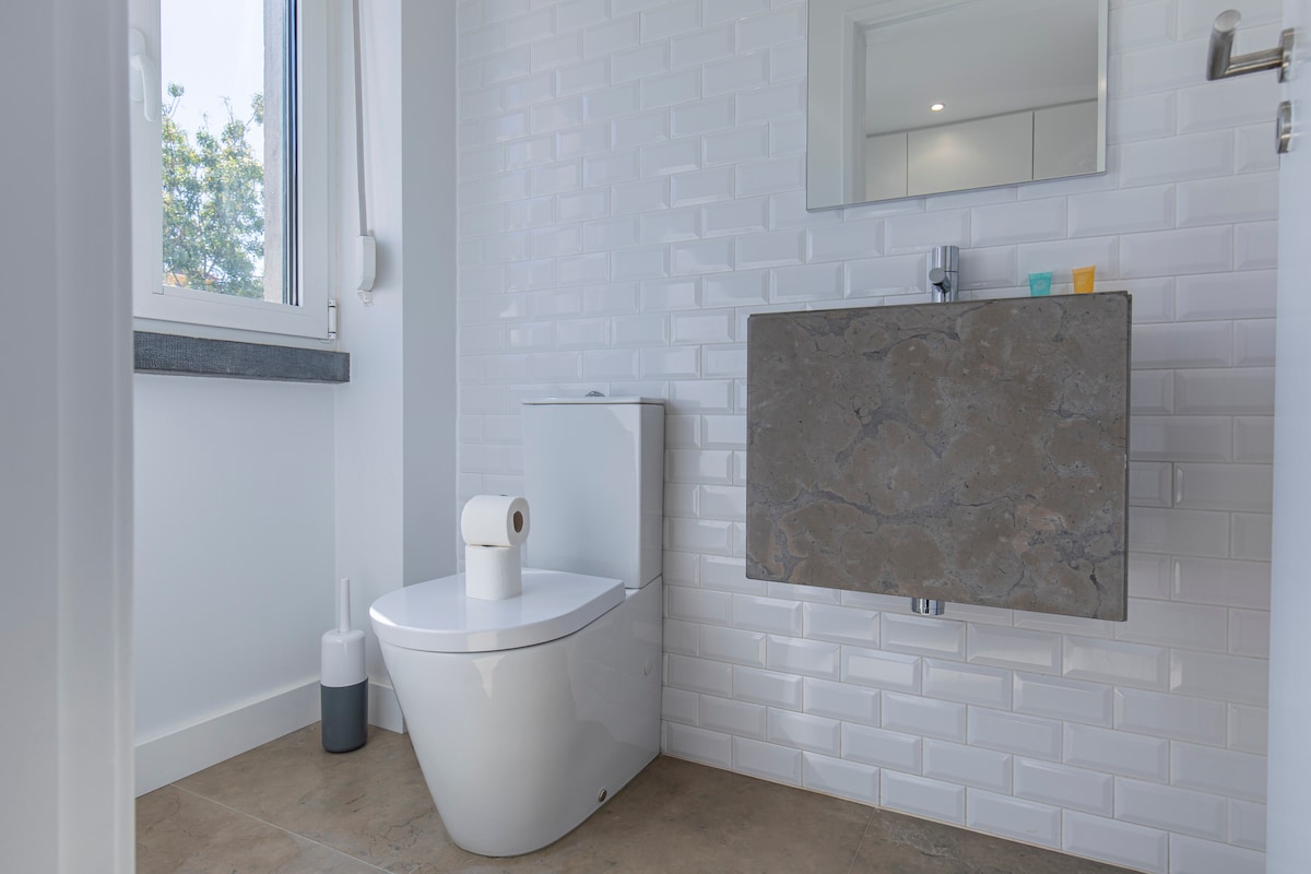 The modern bathroom features a sleek wall-mounted toilet beside a stone vanity. A square mirror reflects natural light, while a window offers views of greenery outside. A minimalist design is complemented by a white tiled wall and neutral-toned flooring.