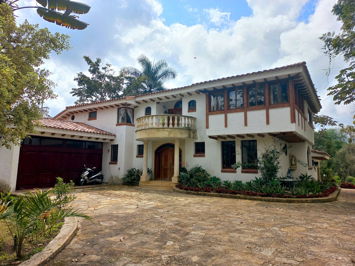A charming two-story house is framed by lush greenery, featuring a decorative balcony on the upper level. The entrance is marked by a wooden door, complemented by large windows that provide natural light to the interior. A stone pathway leads to the main entrance.