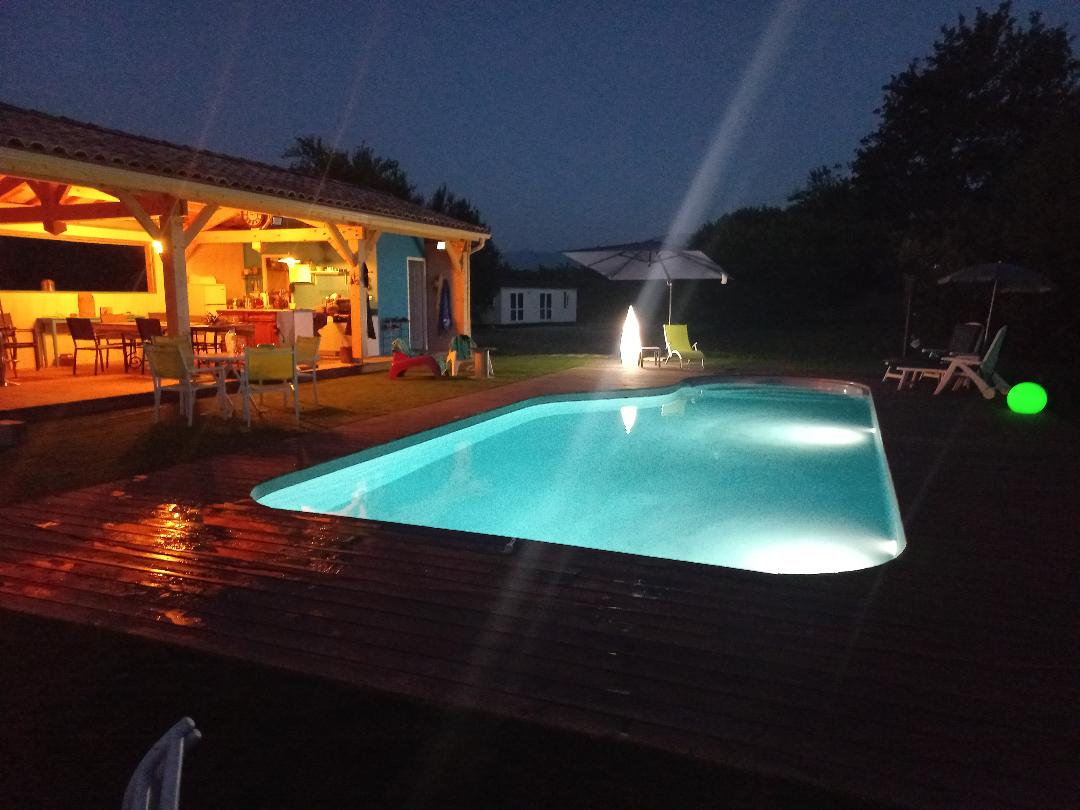 The inviting pool is illuminated by soft lighting, creating a serene atmosphere at night. A wooden deck surrounds the pool, while outdoor seating can be seen under a covered area, providing a comfortable space for relaxation. Umbrellas and lounge chairs enhance the outdoor enjoyment.