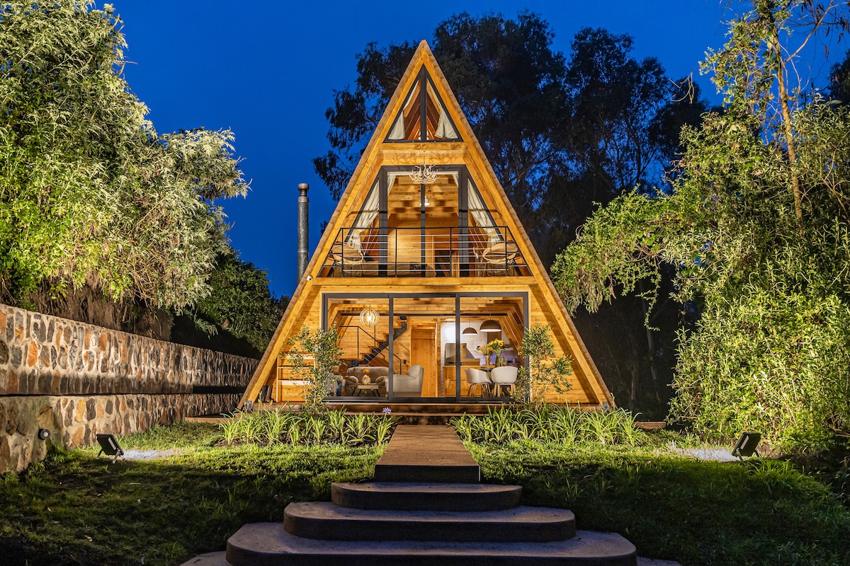 The A-frame chalet is nestled amidst lush greenery, illuminated by soft exterior lighting. Large glass panels showcase the interior's modern design. A stone pathway leads to the entrance, flanked by natural landscaping that enhances the serene outdoor setting.