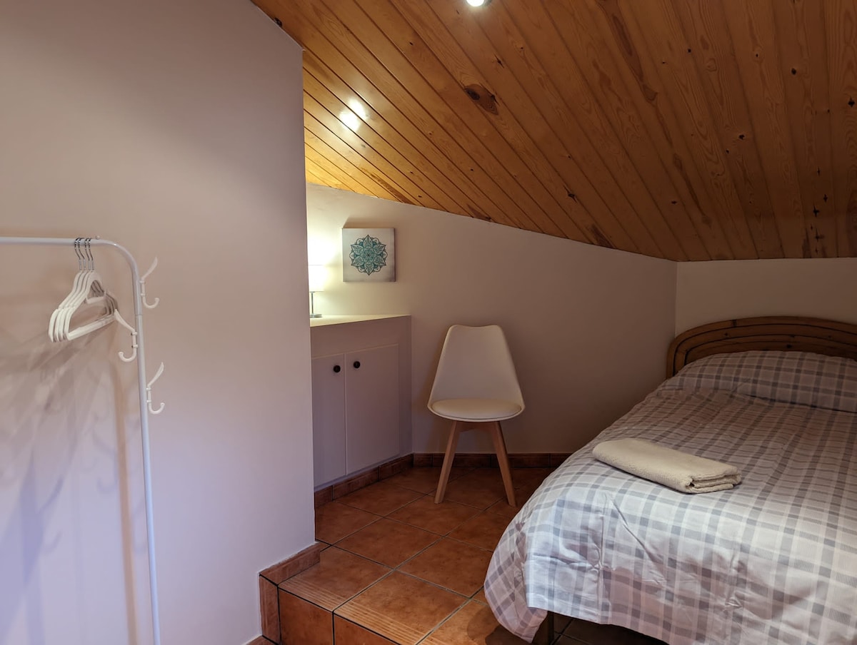 A cozy room features a wooden ceiling and tiled flooring. A spacious bed is positioned against the wall, with a folded towel placed on it. A small white chair and a compact storage unit are present, creating a functional and inviting space.