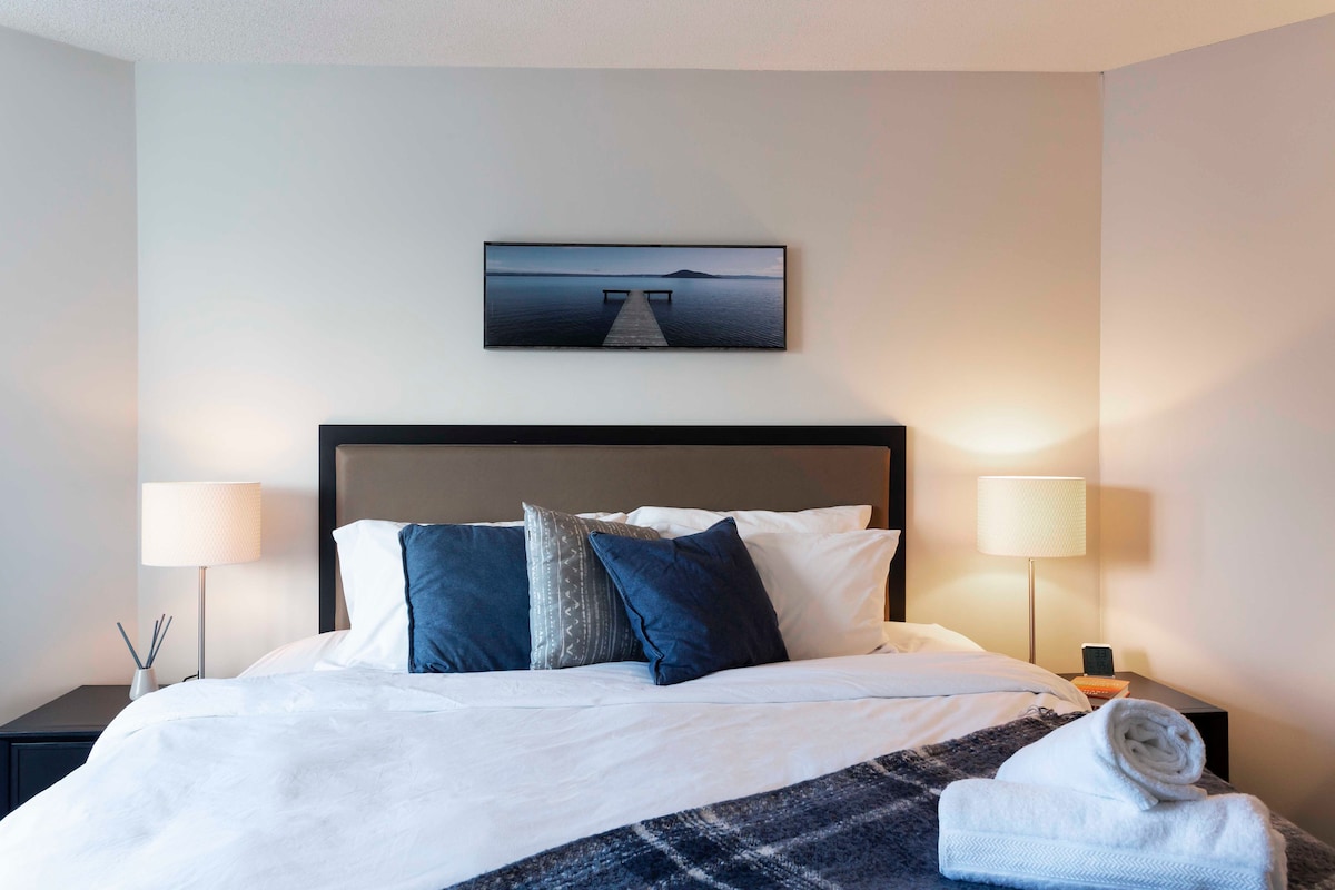 A serene bedroom setting is presented with a spacious bed adorned with soft white linens and plush pillows. Soft lighting from two bedside lamps enhances the calm atmosphere. A minimalist piece of wall art depicting a dock over water adds a tranquil touch to the room.