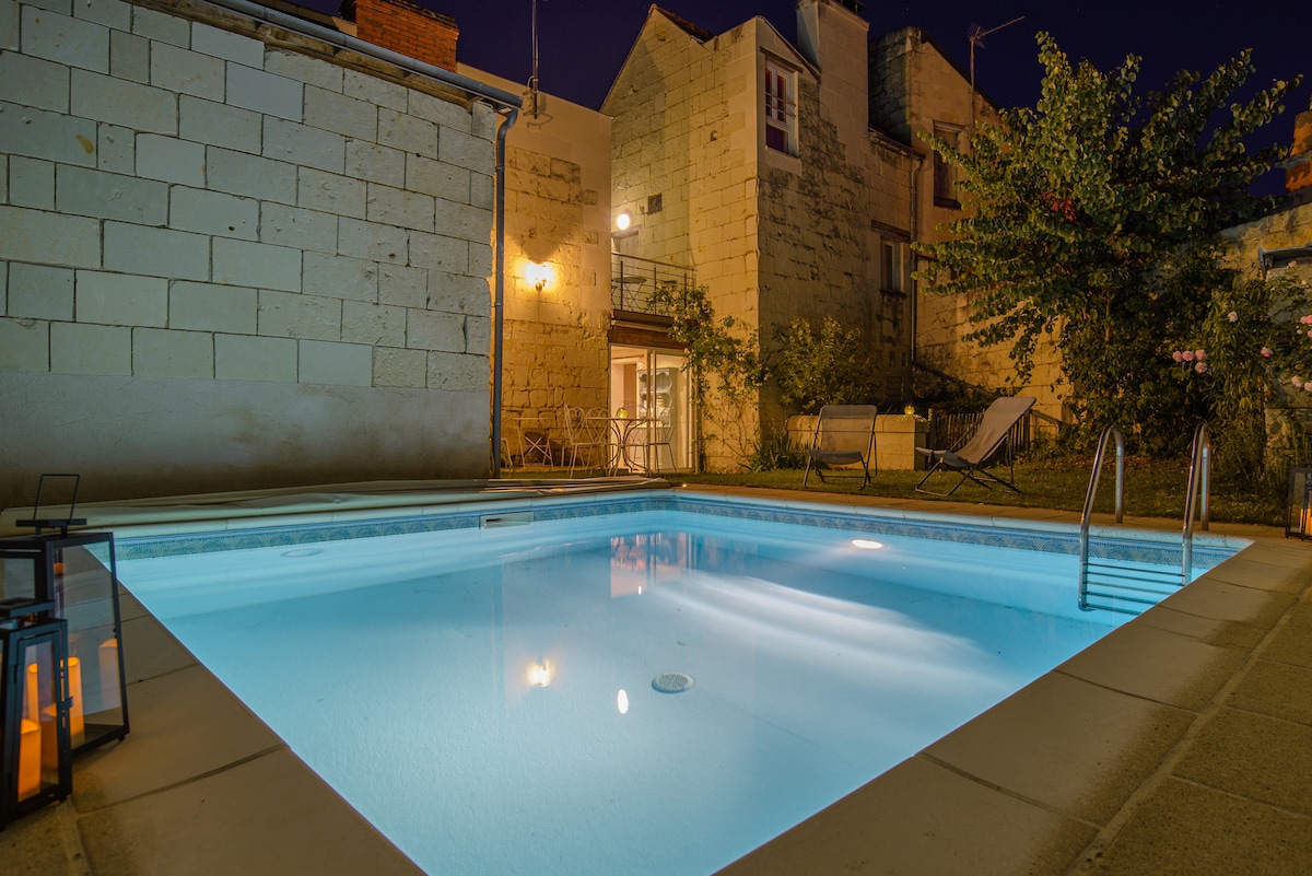 The inviting pool is surrounded by a stone wall, illuminated softly in the evening light. Lounge chairs are positioned nearby, and lush greenery adds a touch of nature to the serene setting. The house is partially visible in the background.