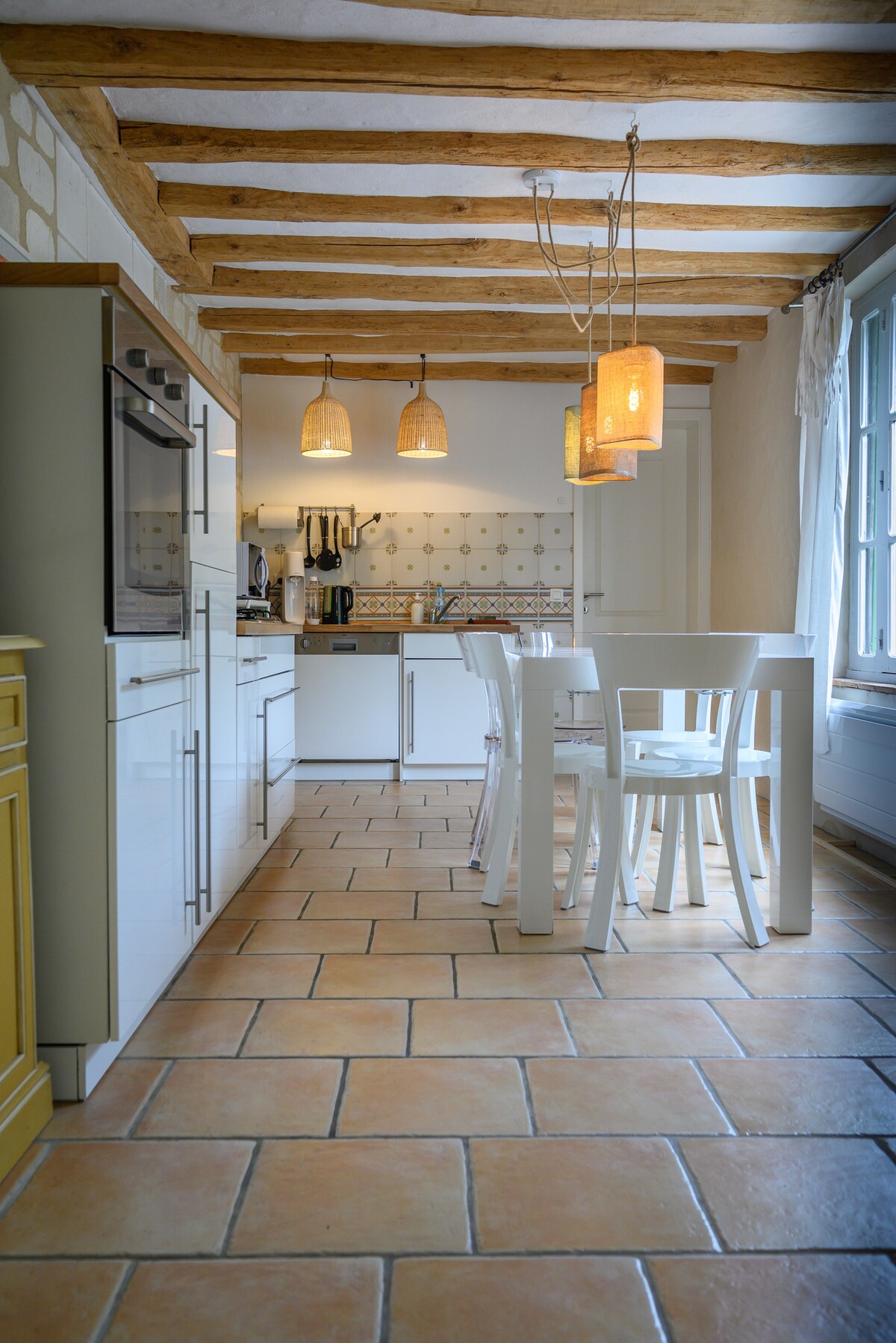 The kitchen features a combination of modern and rustic elements, with sleek white cabinetry and exposed wooden beams. A dining table with four chairs is illuminated by pendant lights, and the tiled floor adds warmth to the space. Appliances are visible against the backdrop of patterned tiles.