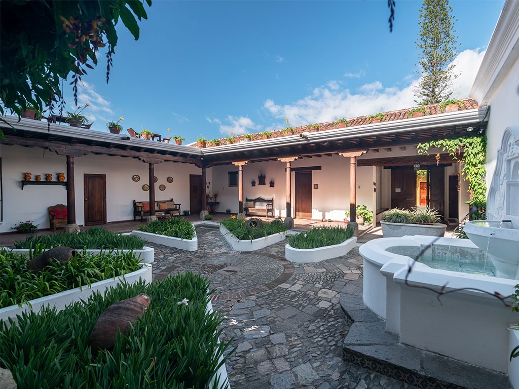 A central courtyard is surrounded by adobe walls, featuring a stone pathway and lush greenery. Wooden doors provide access to the individual spaces, while planters and potted plants add natural beauty. A decorative fountain enhances the serene environment.