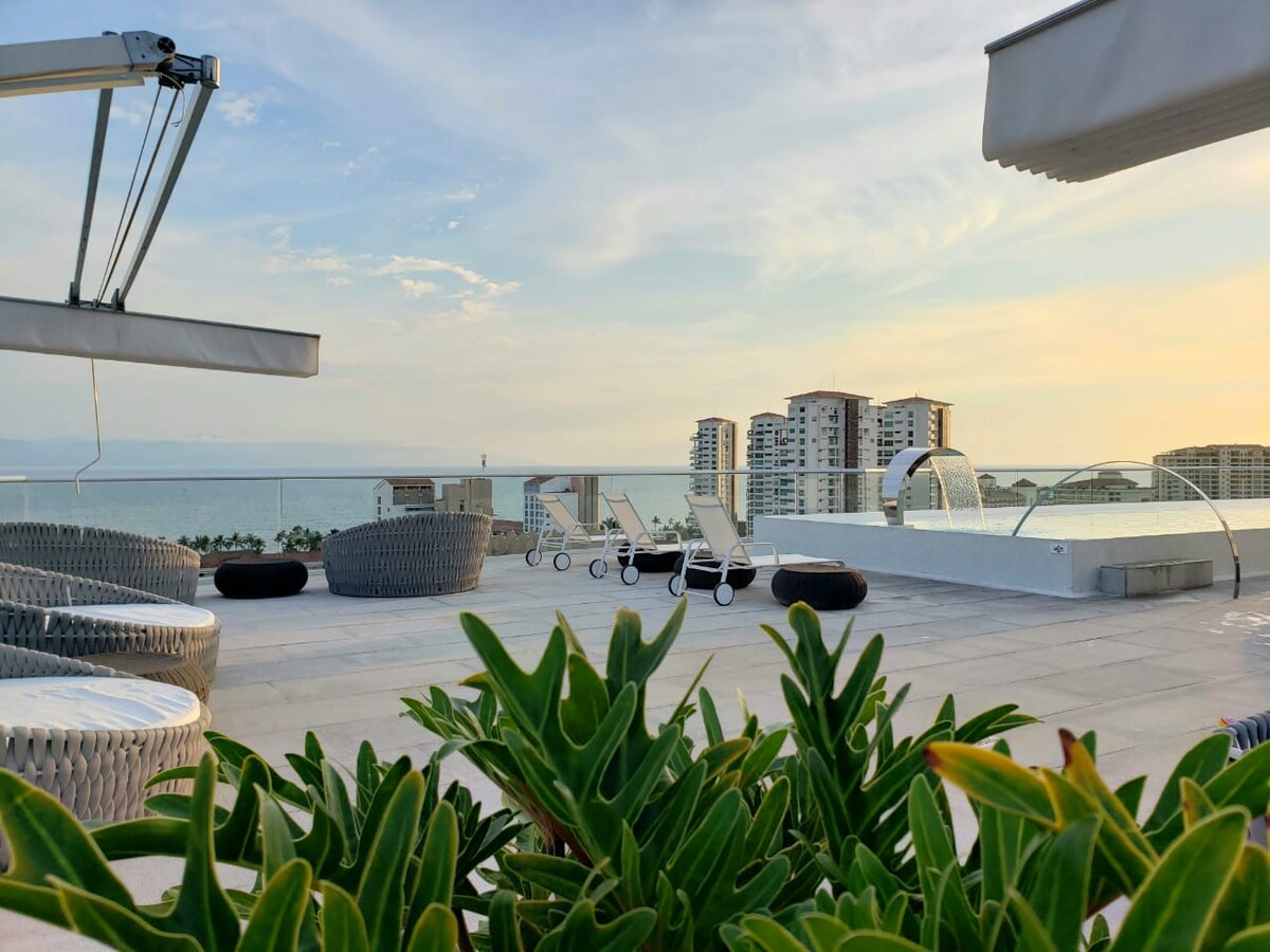 An outdoor terrace offers a scenic view of the ocean and nearby buildings. Comfortable lounge chairs are arranged beside a shimmering pool, with modern seating surrounding the area. Lush green plants frame the scene, enhancing the spacious, relaxing environment.