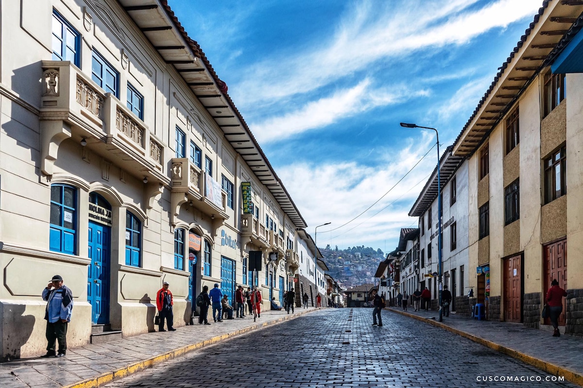 A vibrant street in Cusco is depicted, lined with colonial buildings featuring blue doors and wooden balconies. Cobblestone paving guides pedestrians as they walk along the street. A backdrop of hills reveals the historic charm and lively atmosphere of the area.