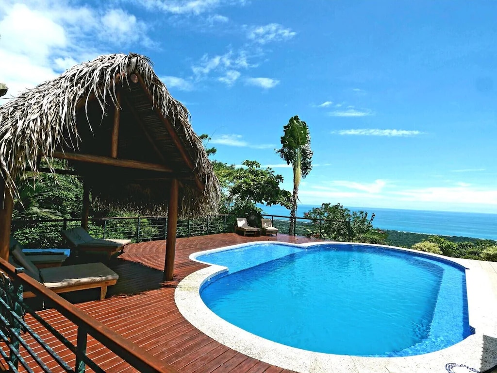 An outdoor deck features a bright blue pool surrounded by lush greenery. A thatched-roof cabana provides shade, with two loungers positioned nearby. The expansive ocean view is visible in the background, set against a clear sky.