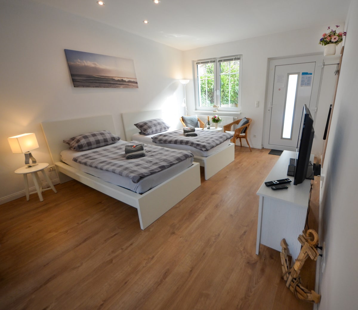 The room features two single beds placed side by side, with a neutral-toned plaid bedding and soft lighting from a bedside lamp. A small wooden table and chair are positioned near the window, allowing natural light to fill the space. A TV is set on a white cabinet, complementing the overall decor.