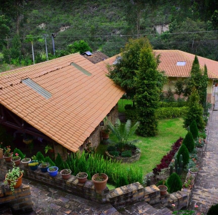 A rustic property is surrounded by lush greenery, featuring terracotta roofs and a well-maintained garden. Potted plants line the path, enhancing the outdoor space. The arrangement of trees and shrubs offers a natural, serene environment.