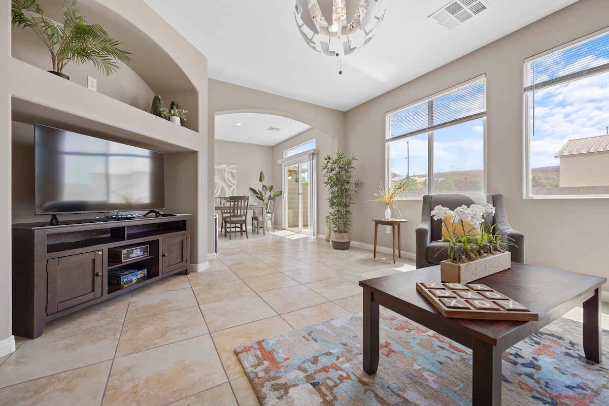 A modern living area is highlighted with natural light from large windows. A dark wooden coffee table sits atop a textured rug, complemented by a comfortable armchair and a television unit. Potted plants add greenery, creating a welcoming ambiance.