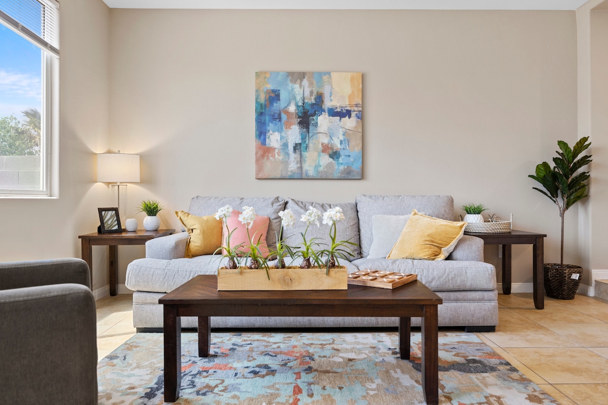 A welcoming living area is highlighted by a light gray sofa adorned with colorful cushions. A wooden coffee table sits centrally, featuring a planter with white flowers. Soft natural light enters through a nearby window, and an abstract wall art piece adds a touch of color.