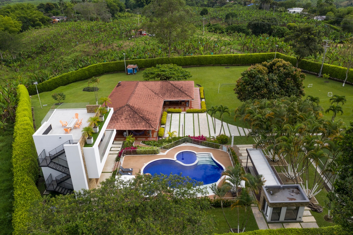 The property is set within a spacious, green landscape, featuring a pool and multiple outdoor areas. A well-maintained garden surrounds the home, while ample seating is available on a terrace. The building showcases a mix of modern and traditional architectural elements.